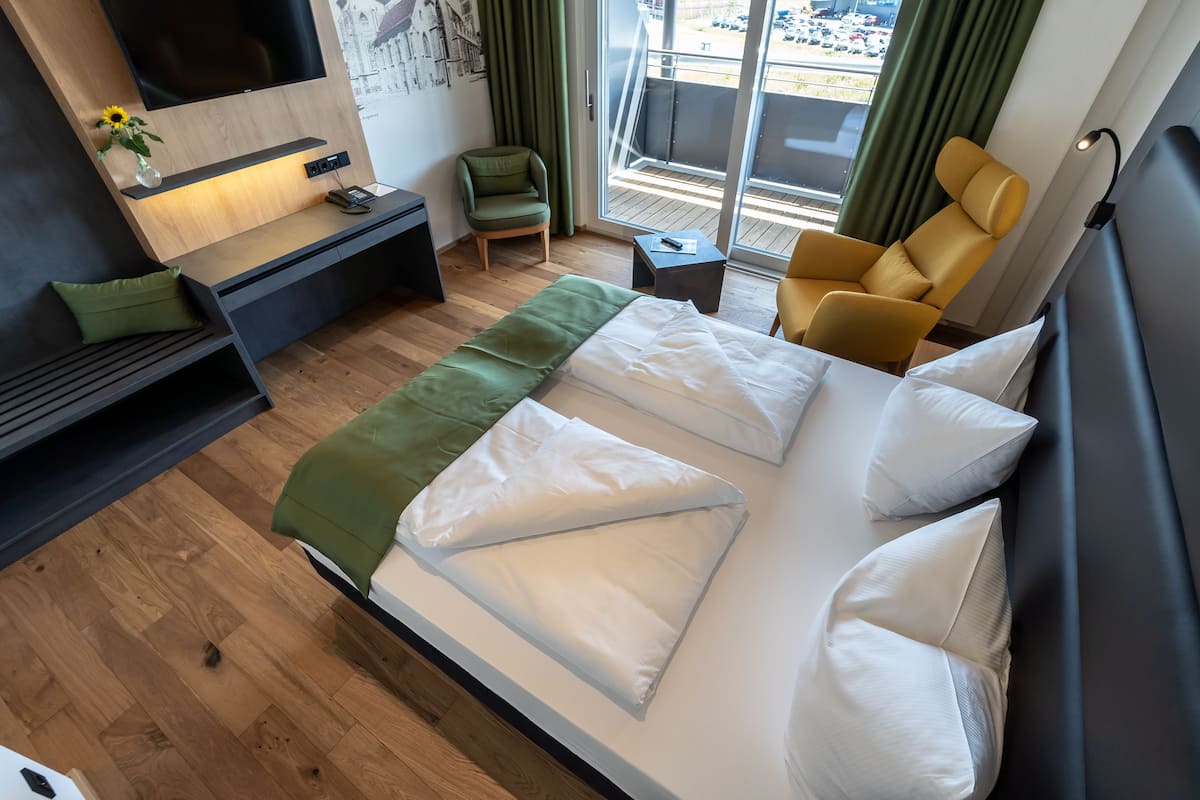 Une chambre d’hôtel moderne dotée d’un grand lit, d’un bureau avec une lampe, d’une chaise, d’une petite table et offrant une vue sur la ville grâce à de grandes fenêtres.
