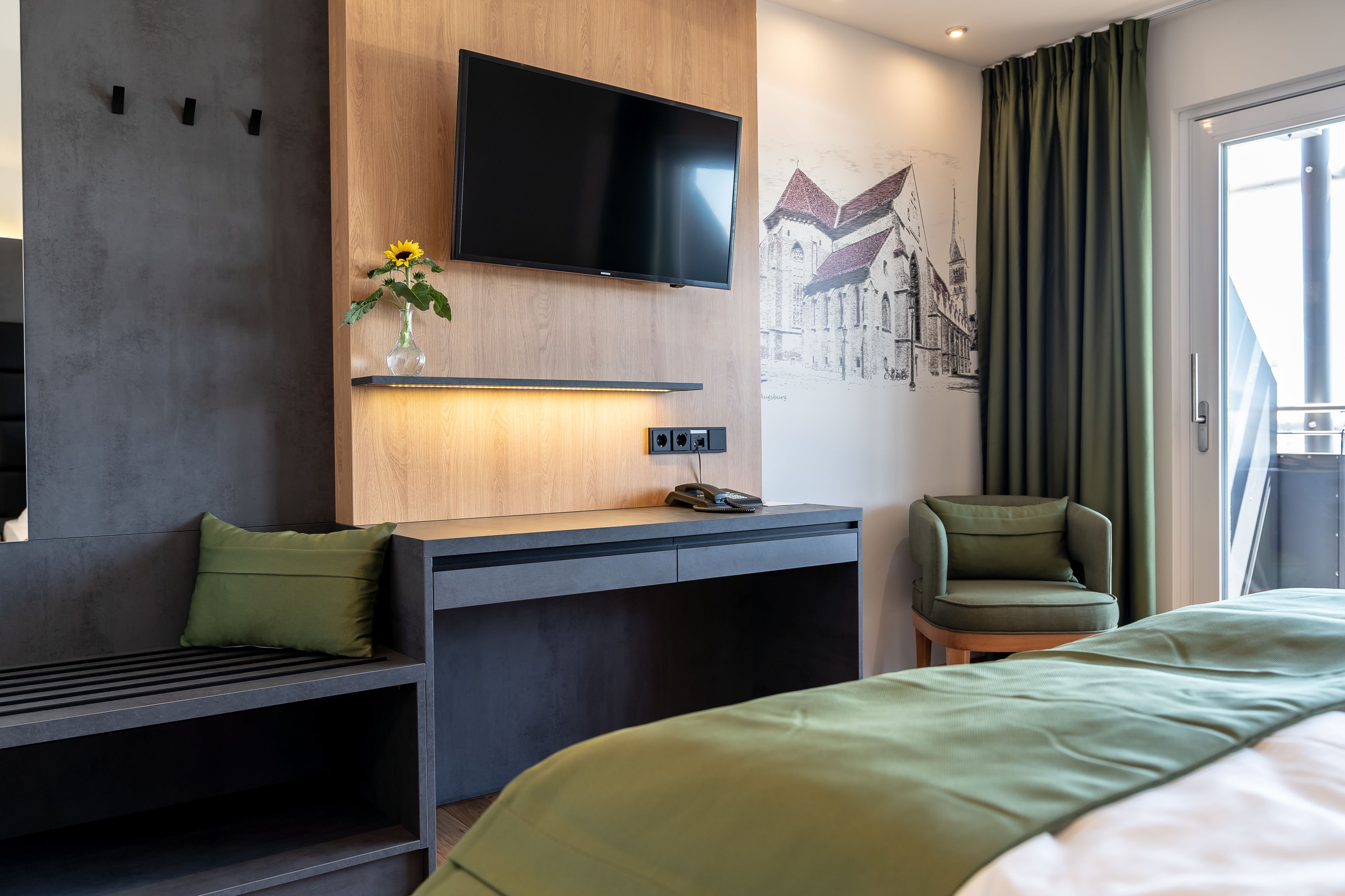 Une chambre d’hôtel moderne équipée d’un lit, d’un bureau avec un téléviseur à écran plat, d’une chaise et d’une étagère murale agrémentée d’un vase de fleurs.