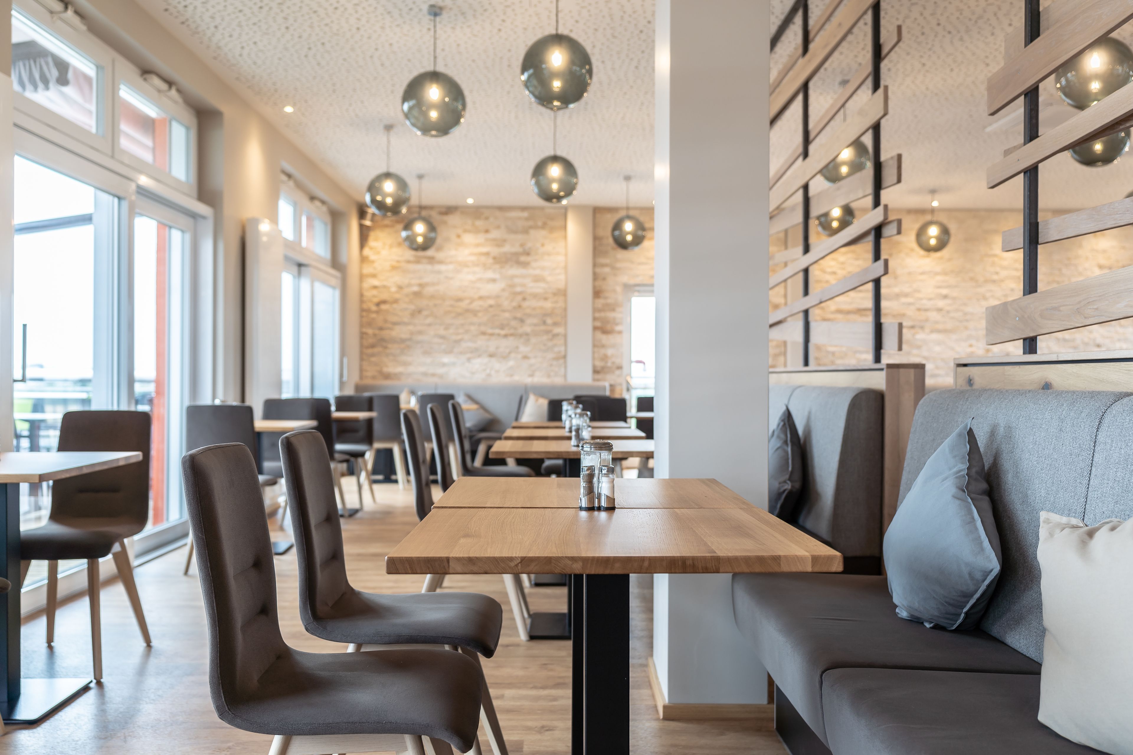 A modern dining area with wooden tables and chairs, a sofa with cushions, and pendant lighting.