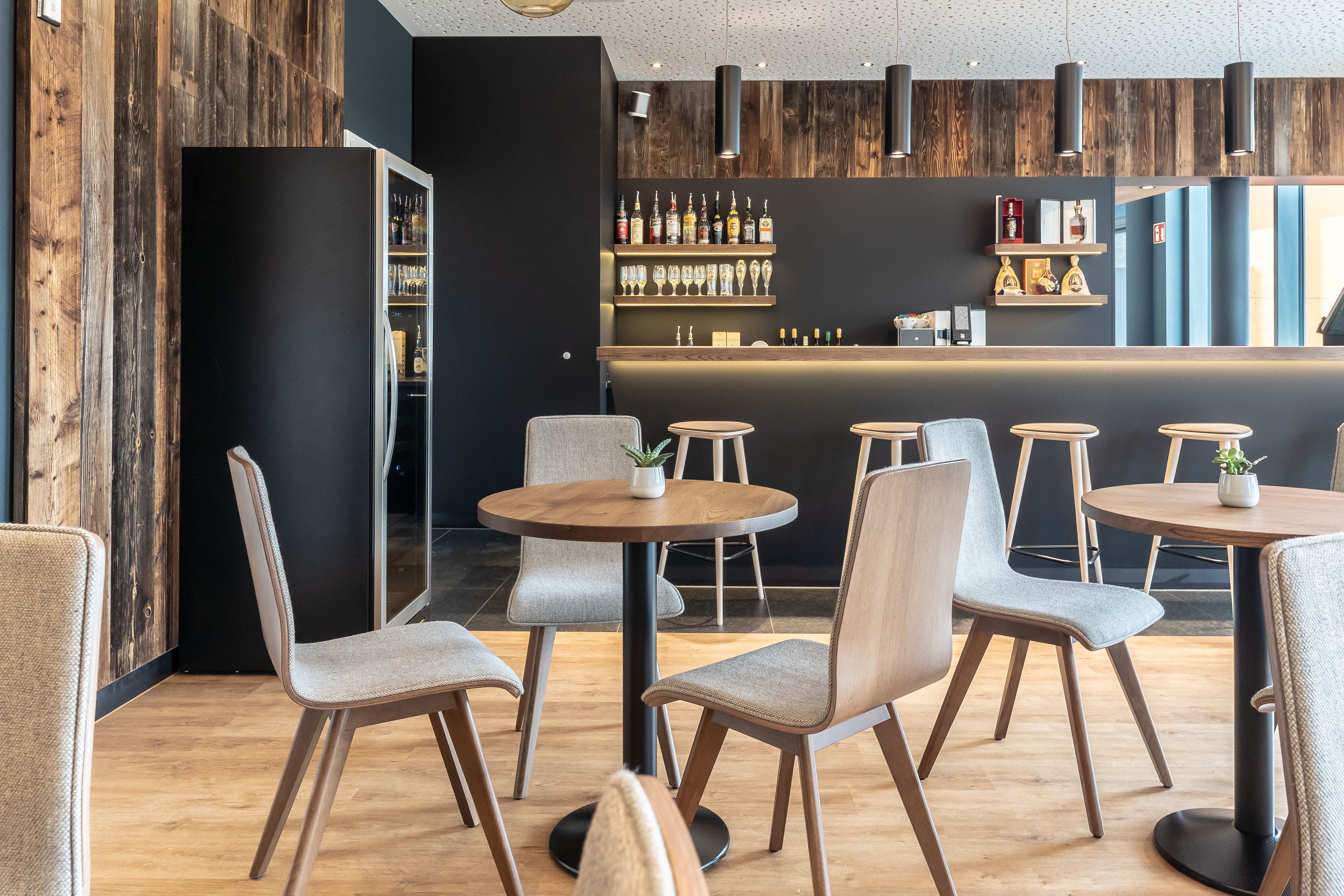Un área de bar moderna con mesas y sillas redondas, suelo de madera y una barra con estantes llenos de botellas y vasos.