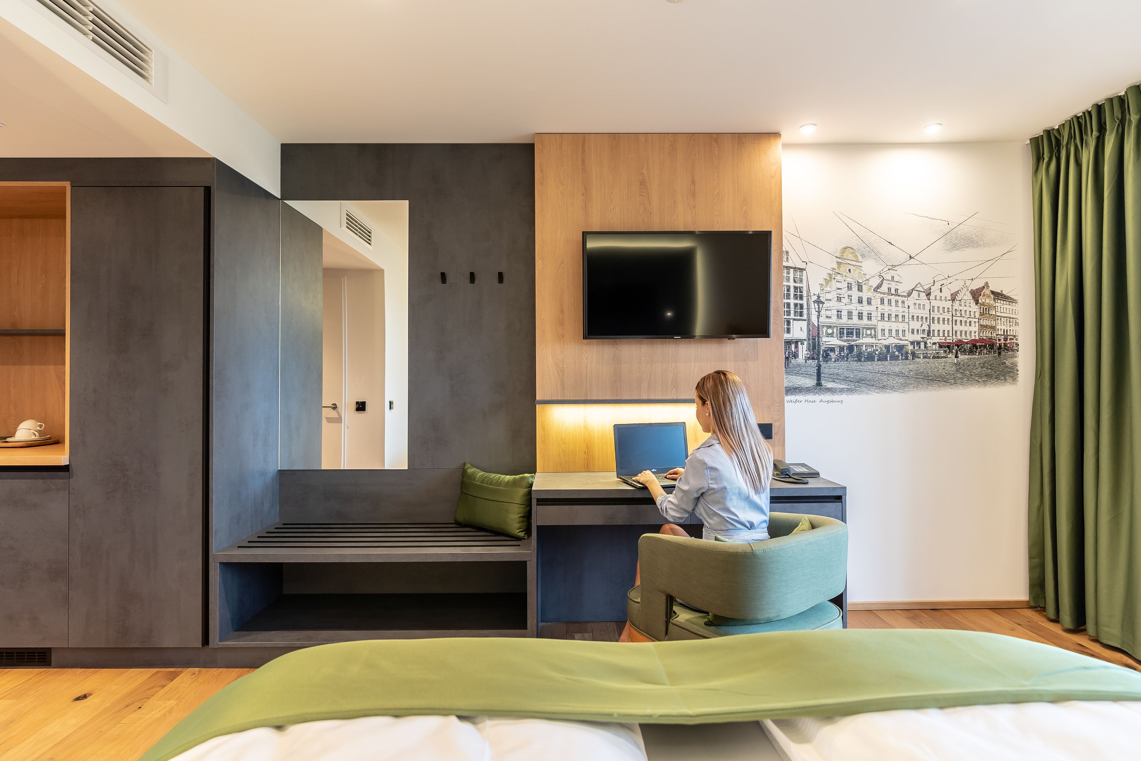 Une chambre d’hôtel moderne, équipée d’un lit, d’un bureau avec un ordinateur portable, d’une télévision et d’un tableau représentant un paysage urbain accroché au mur.
