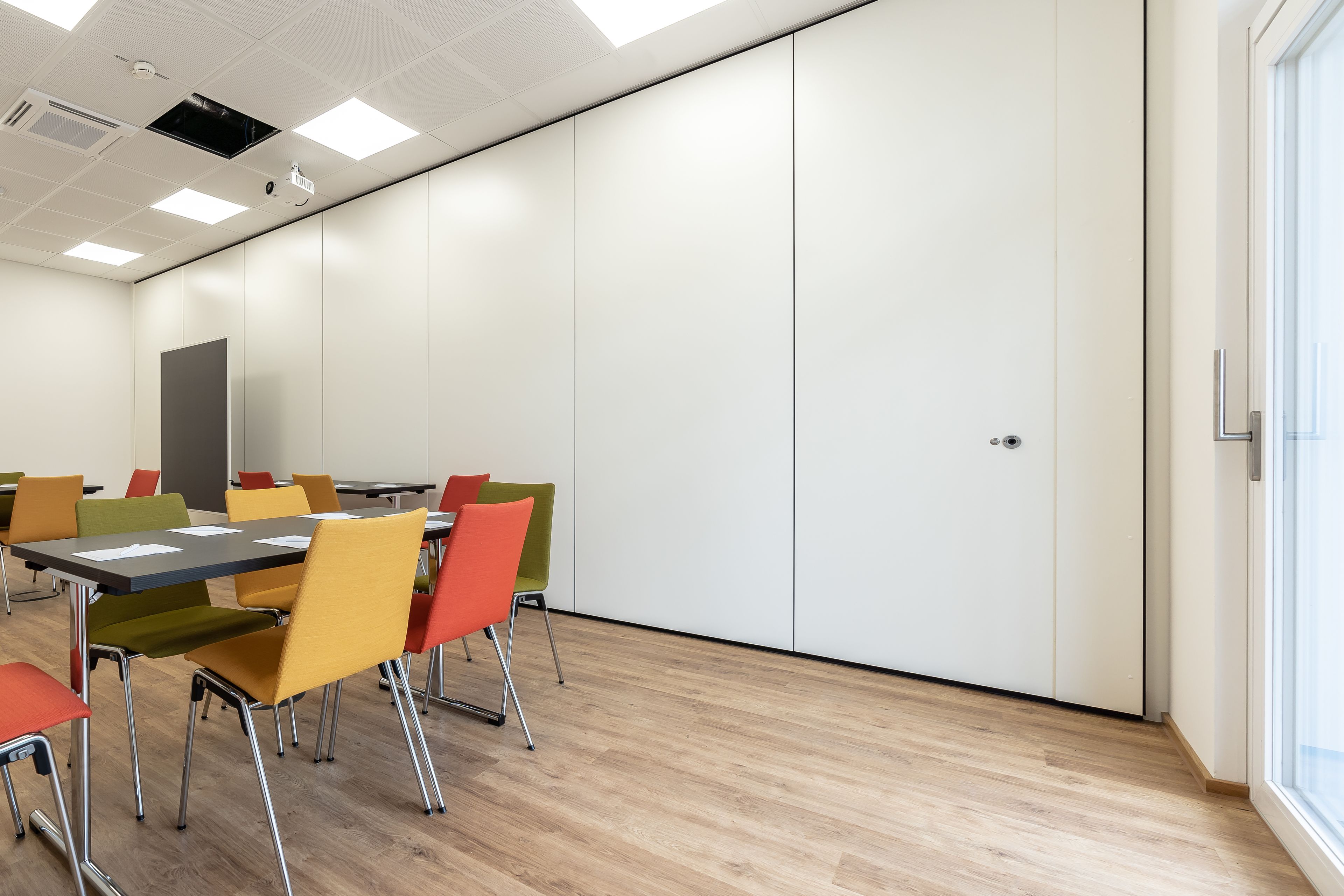 A conference room with a long table, colorful chairs, white walls, and a door.