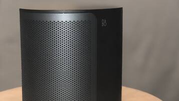 Un haut-parleur noir posé sur une table en bois, avec un lit et des oreillers en arrière-plan.