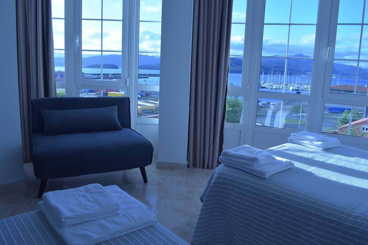 Habitación de hotel con cama, sofá y vistas a un puerto deportivo y montañas.