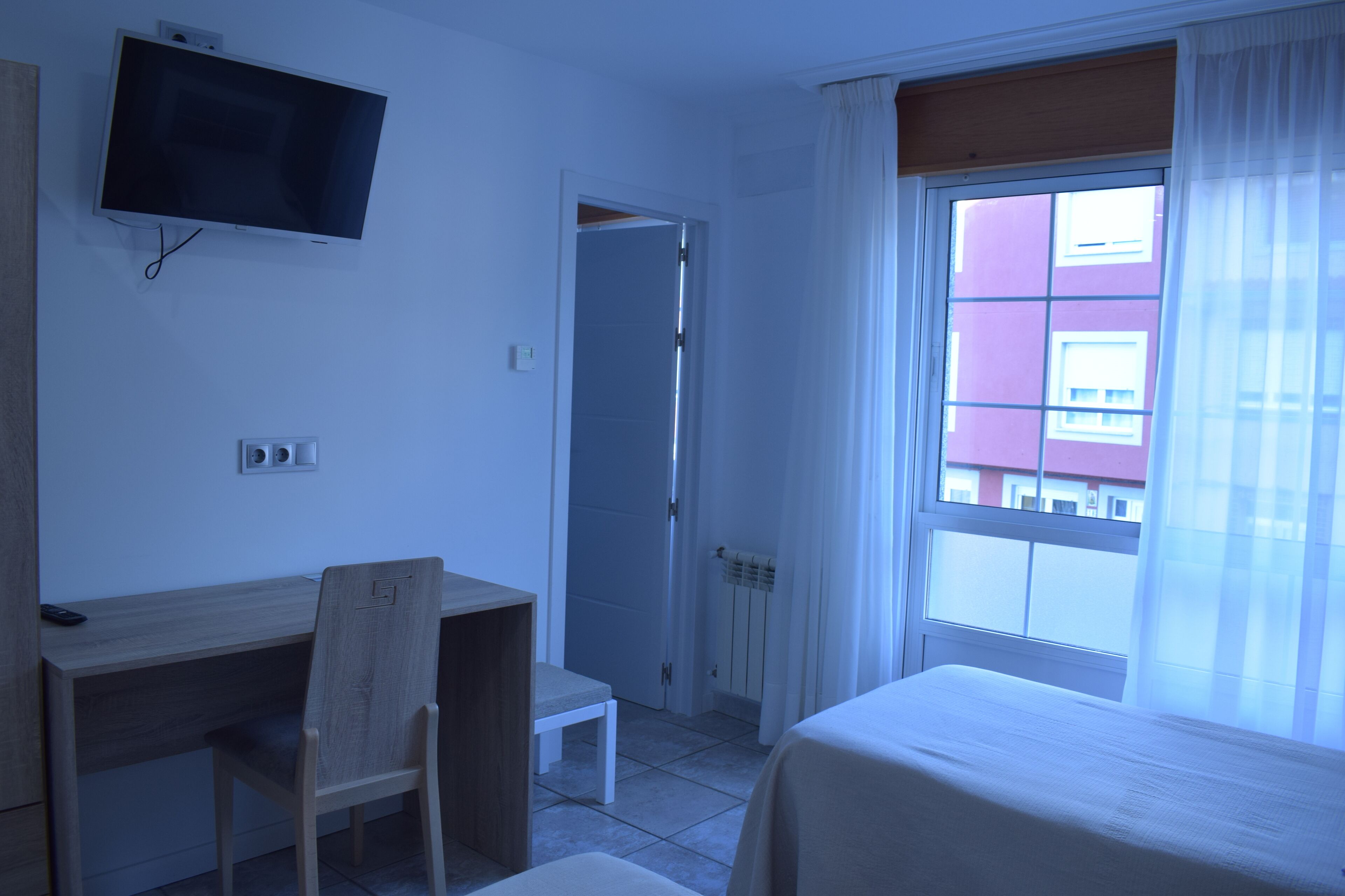A hotel room with a bed, a desk, a chair, a television mounted on the wall, and a window with curtains.