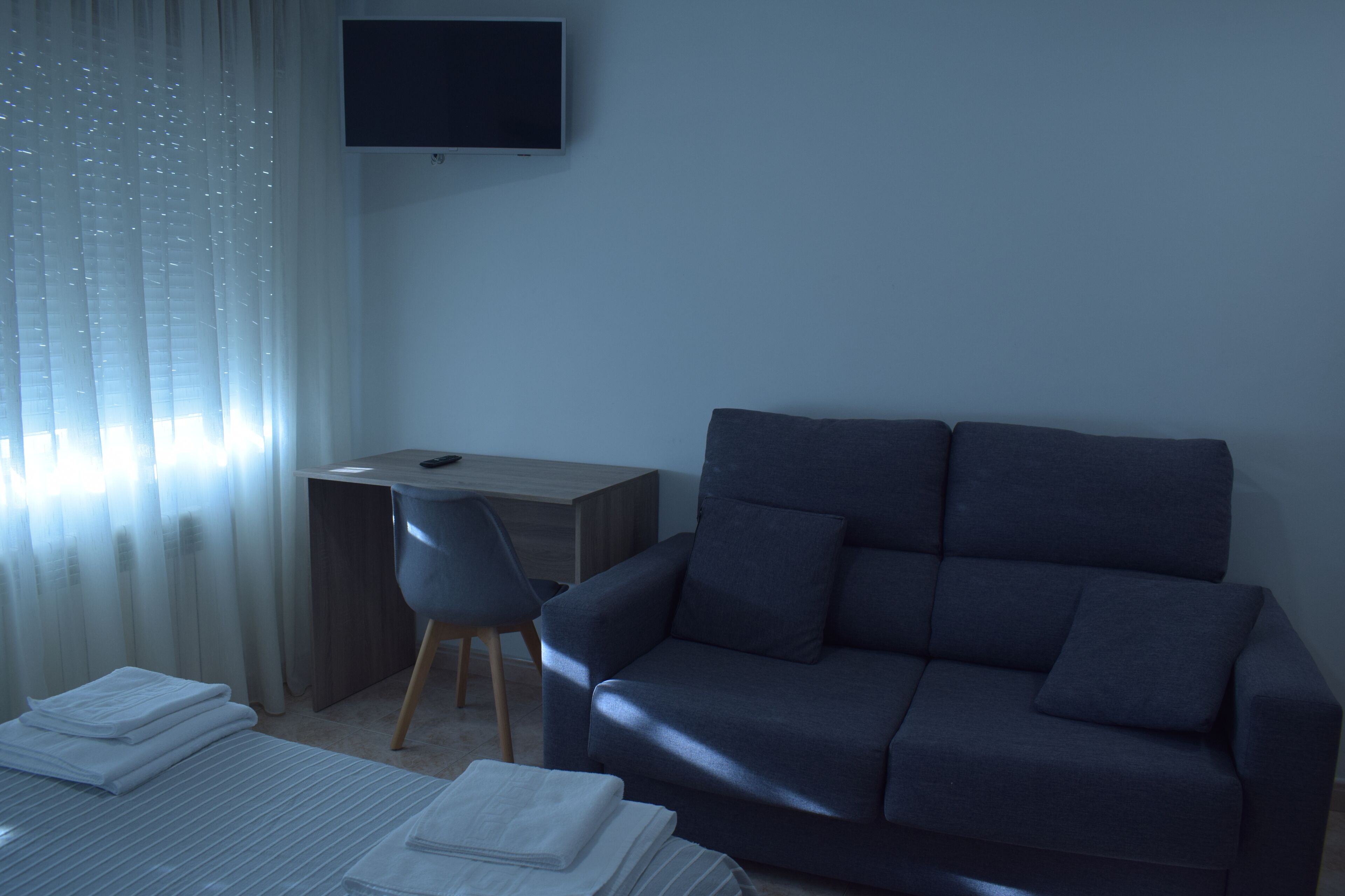 A hotel room with a dark gray sofa, a wooden desk, a chair, and a TV mounted on the wall.