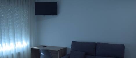 A hotel room with a dark gray sofa, a wooden desk, a chair, and a TV mounted on the wall.