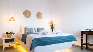 Un lit bien fait, recouvert d’une courtepointe turquoise, avec deux oreillers blancs et une serviette enroulée. Deux chapeaux sont accrochés au mur, il y a une petite table de chevet avec une plante, et une table de nuit en bois.