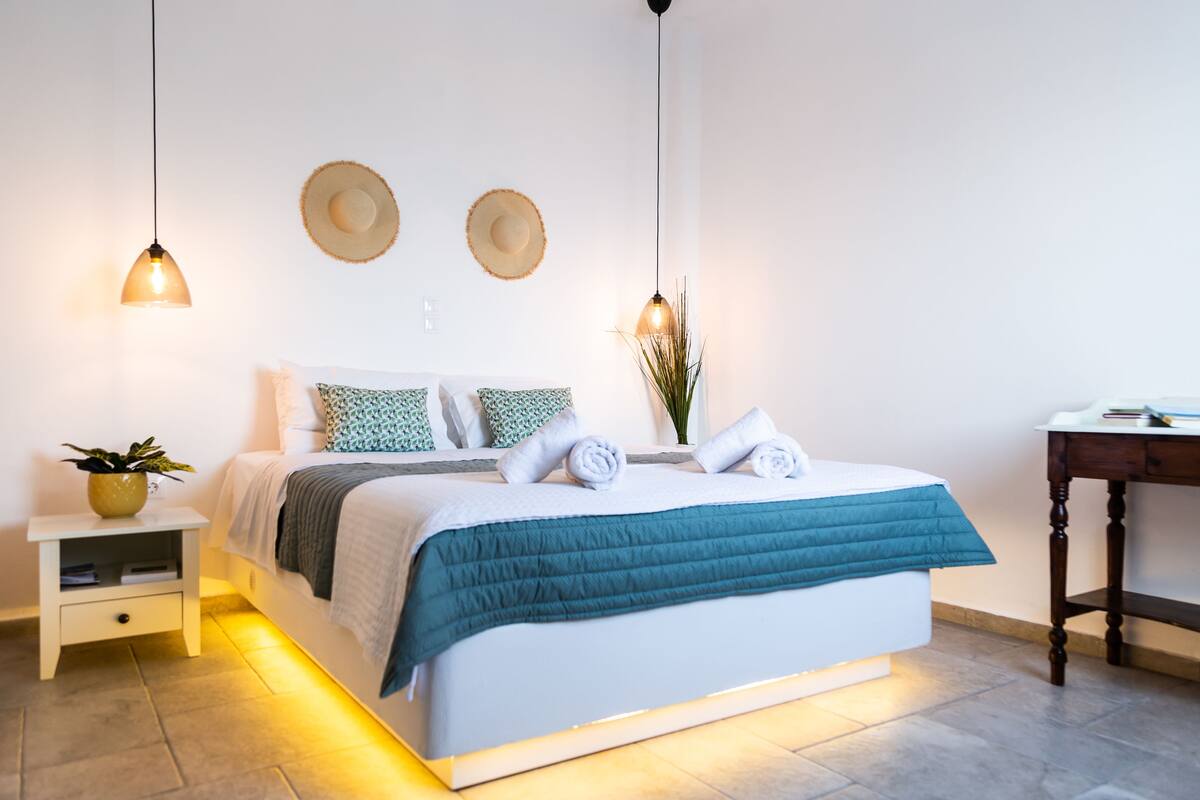Una cama bien tendida con un edredón verde azulado, dos almohadas blancas y una toalla enrollada. Se observan dos sombreros colgados en la pared, una mesita de noche con una planta y un buró de madera.