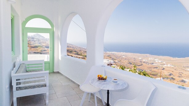 Un balcon avec un banc et une table blancs, une porte verte et une vue sur la mer et les collines.