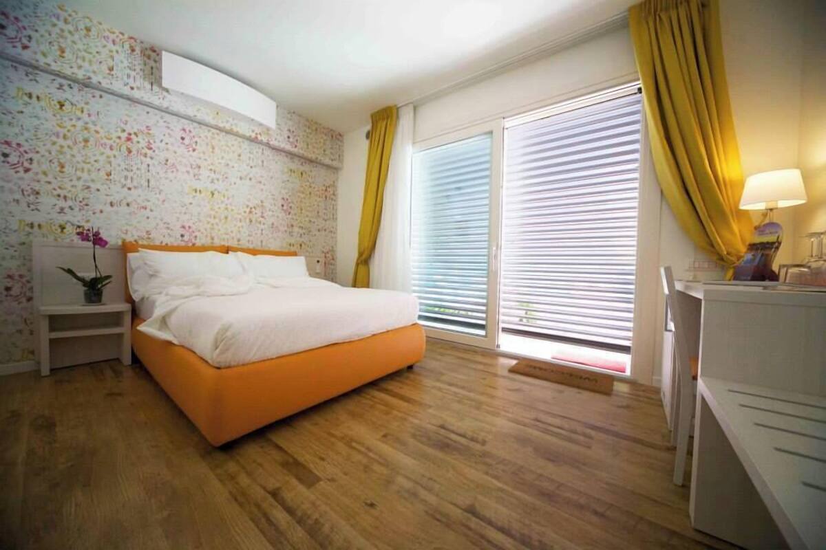 Ein Schlafzimmer mit Holzboden, einem Bett mit weißer Bettwäsche, einem Nachttisch mit Pflanze, einem Schreibtisch mit Lampe und einem Fenster mit gelben Vorhängen.