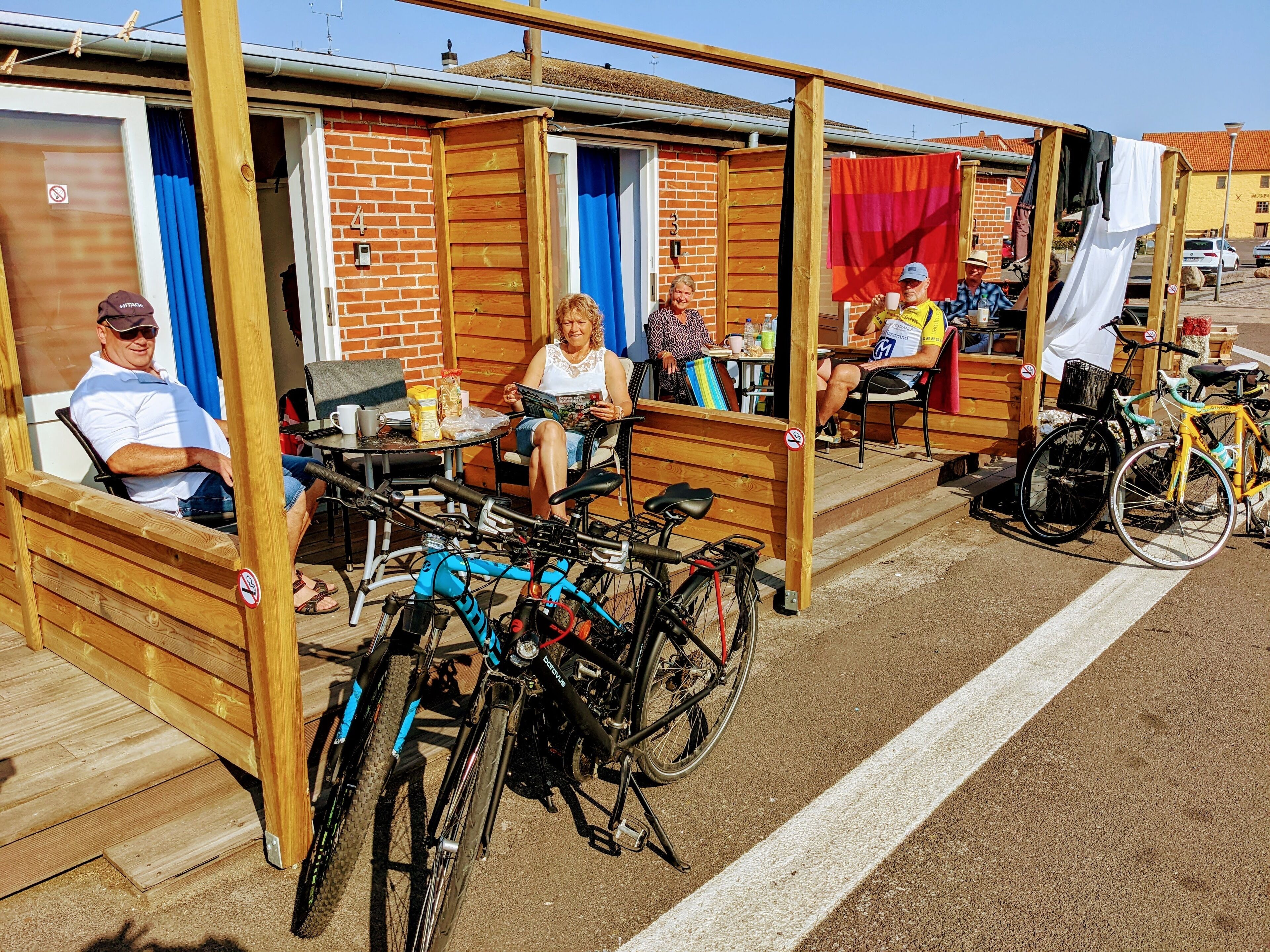 A covered outdoor seating area with wooden benches and tables, bicycles parked nearby, and a brick building in the background.