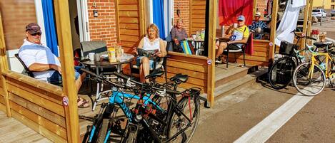 A covered outdoor seating area with wooden benches and tables, bicycles parked nearby, and a brick building in the background.