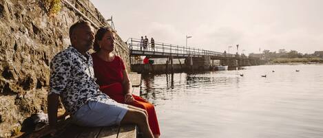 A couple sitting on a wooden dock by a calm river, with a bridge and people in the background.