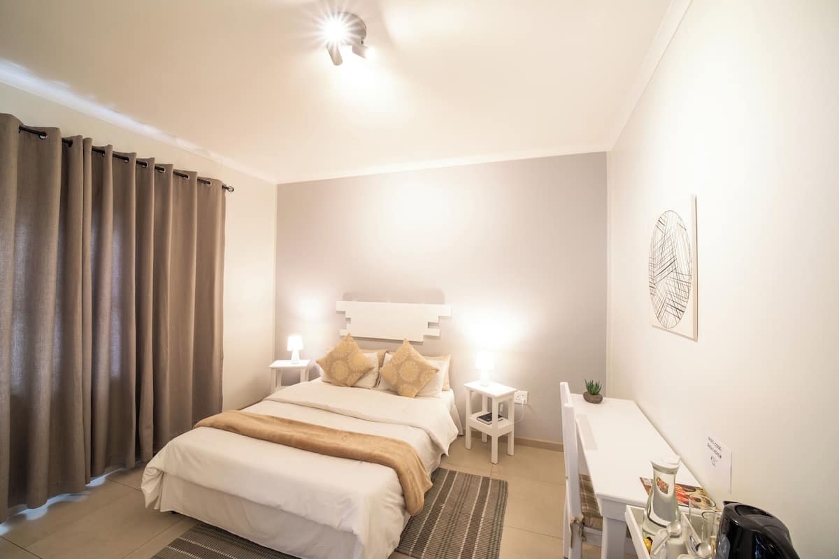 Hotellihuone, jossa on sänky, yöpöydät, työpöytä, tuoli, pieni kasvi ja seinätaideteos.