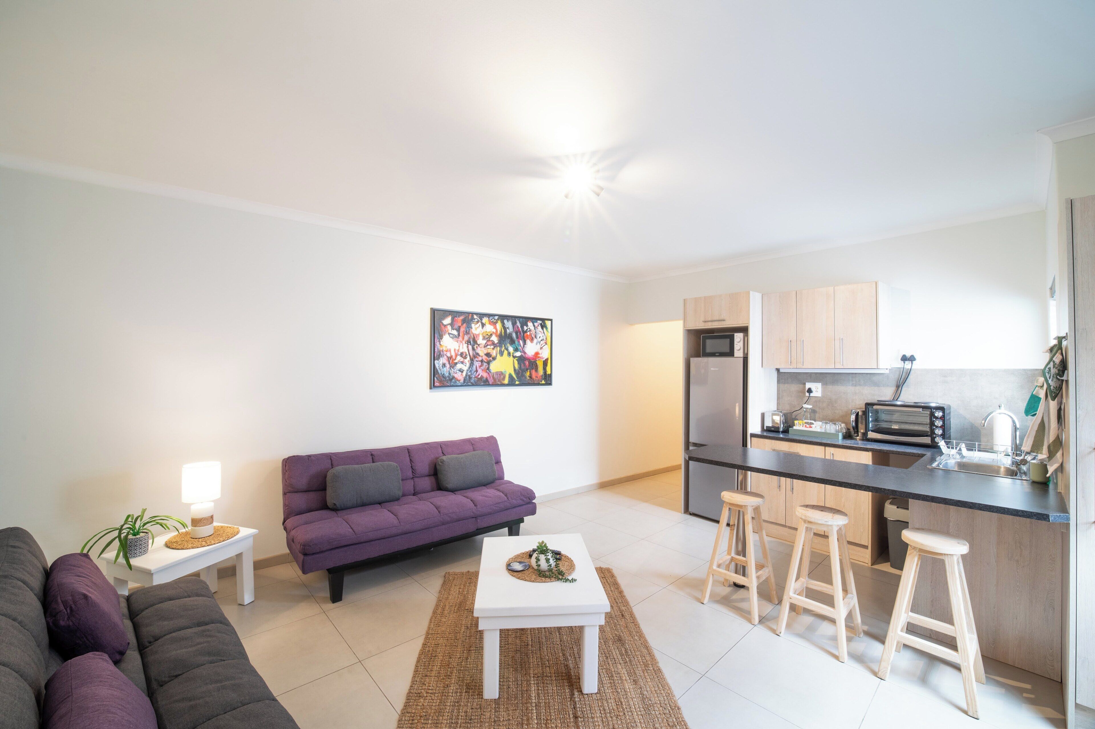 A modern apartment with a living room, purple sofa, white coffee table, kitchen area with a refrigerator, and a bar stool.