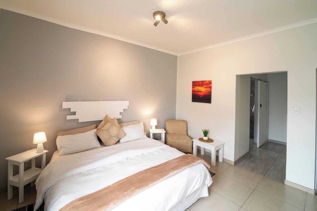 Moderni hotellihuone, jossa on suuri sänky, yöpöydät lamppuineen, tuoli, pieni pöytä, jossa on kasvi, auringonlaskua kuvaava taulu ja ovi, joka johtaa toiseen huoneeseen.