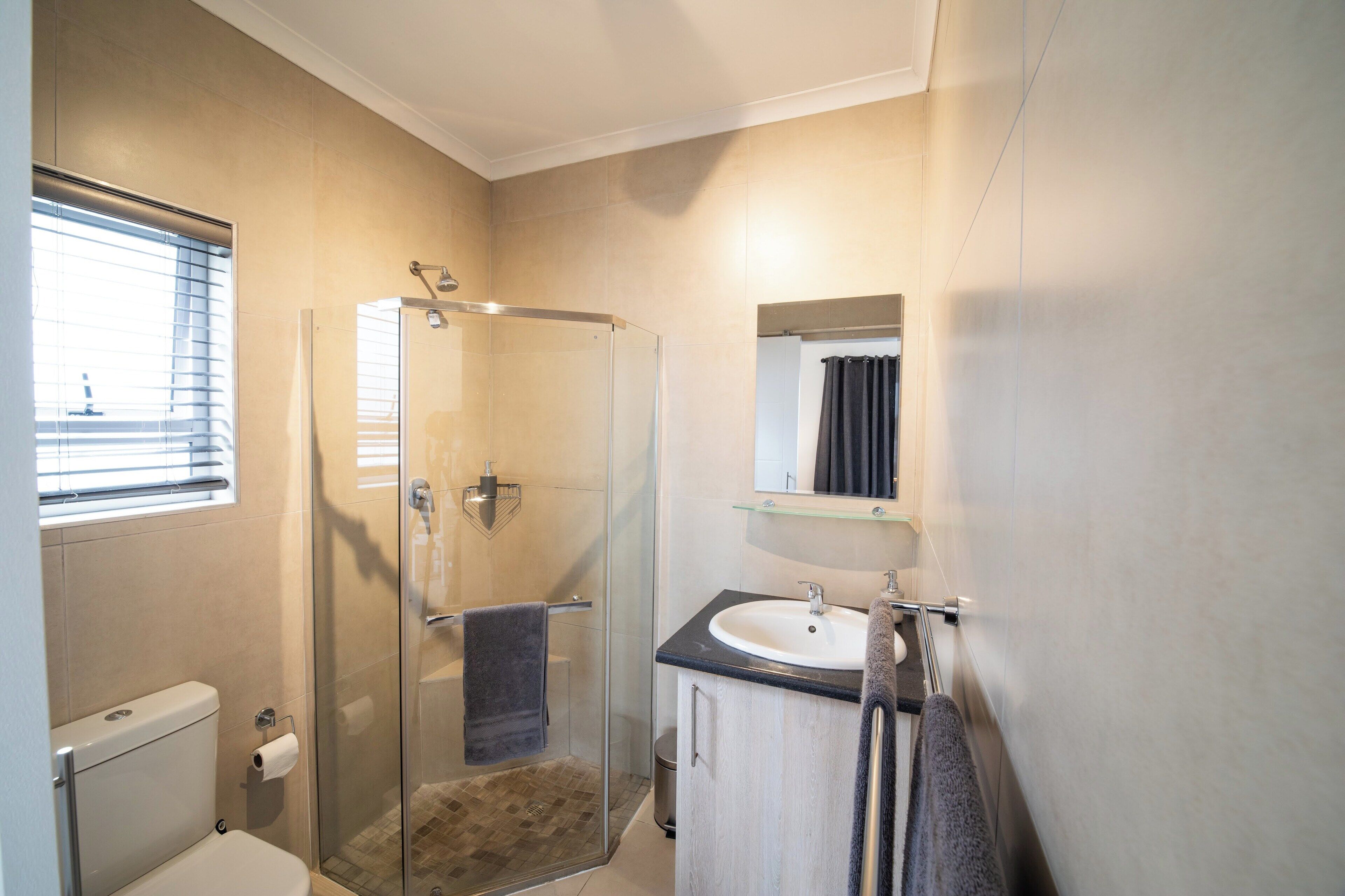 A bathroom with a glass-enclosed shower, a toilet, a sink with a mirror above it, and a window with blinds.