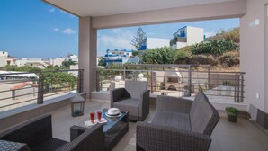 A balcony with wicker furniture, a table with drinks, and a view of buildings and greenery. - Gras Houses Agia Pelagia (Malevizi)