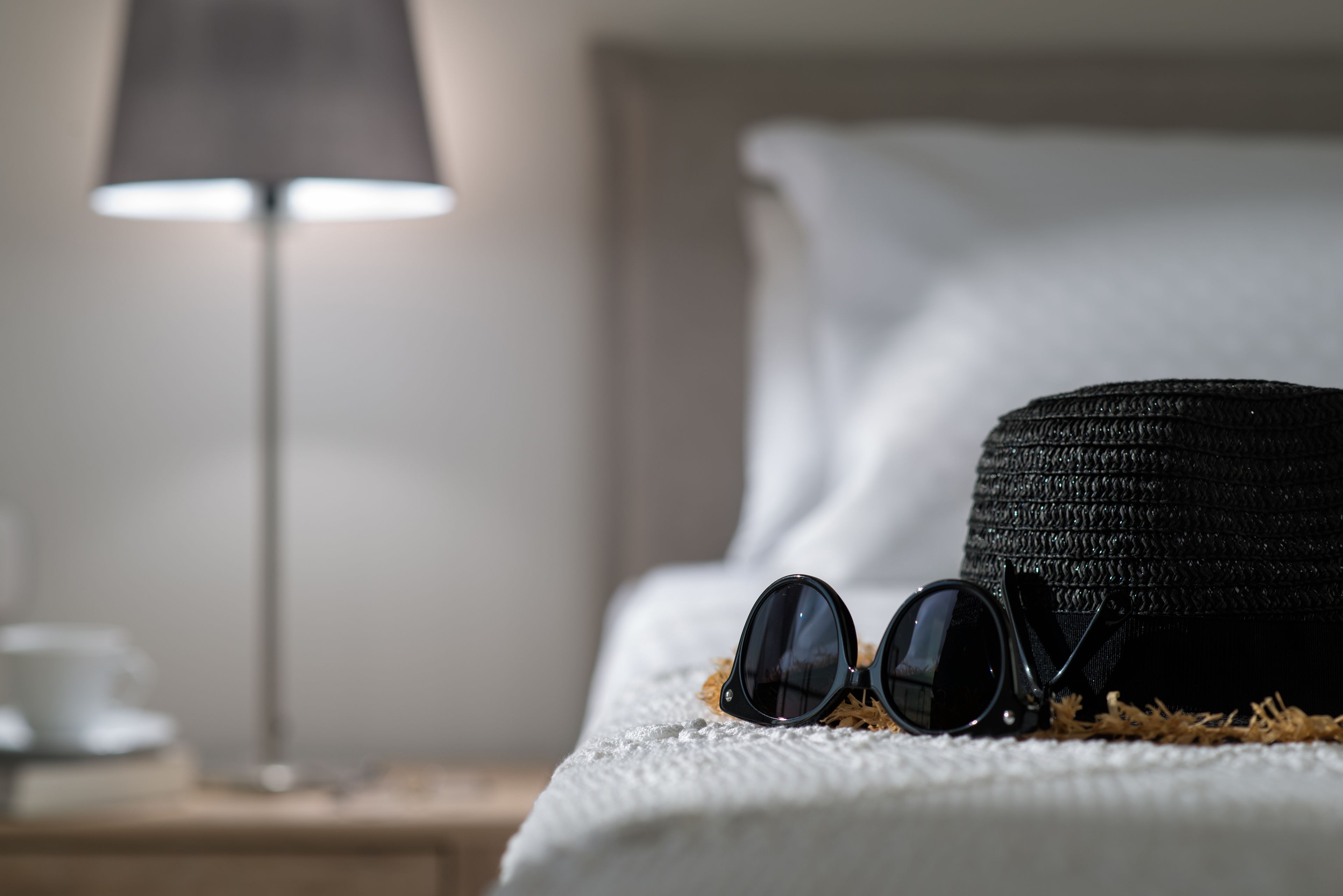 Ein Bett mit weißer Decke, einem schwarzen Hut und einer Sonnenbrille auf einer strukturierten Oberfläche.