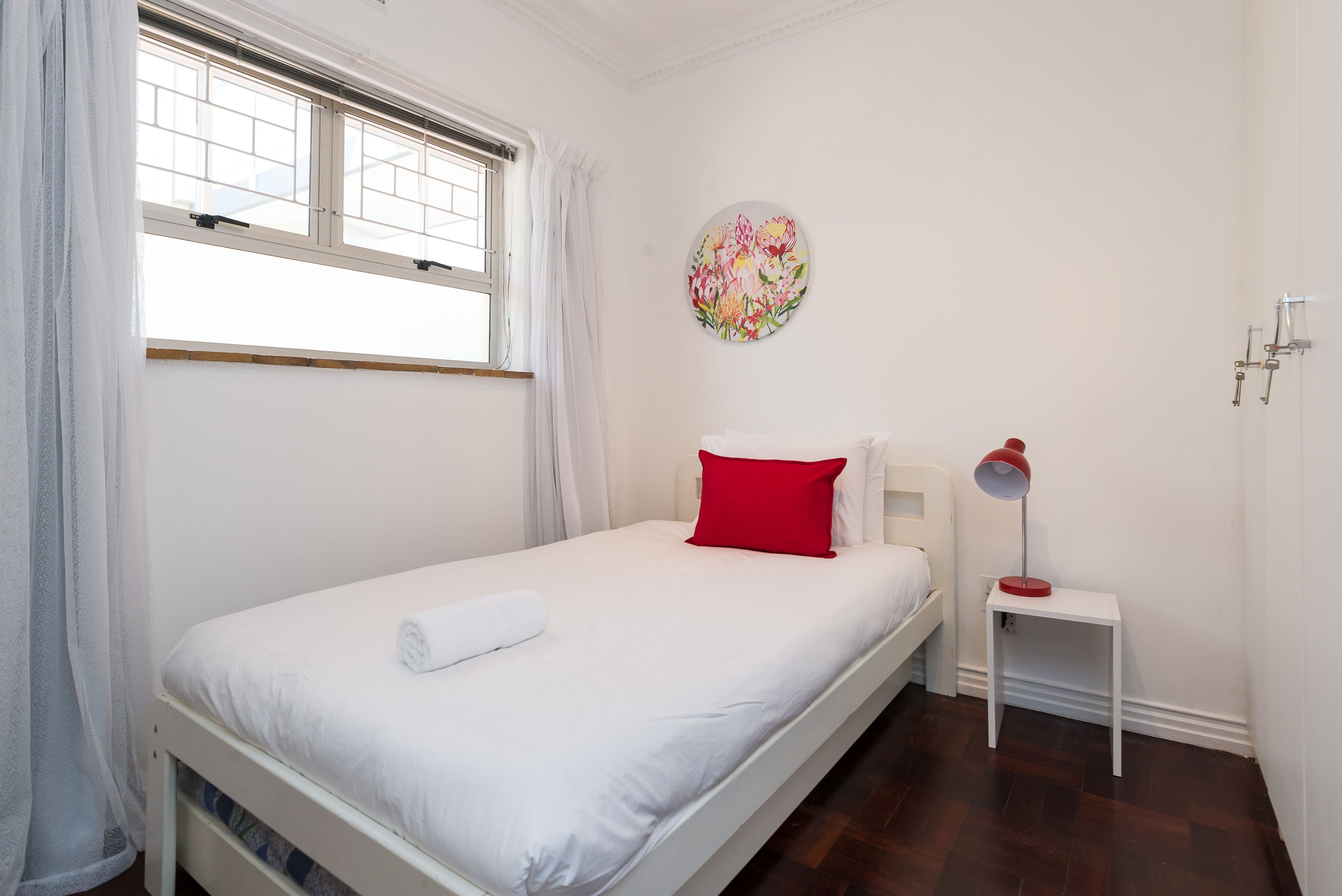 Een enkel bed met wit beddengoed en een rood kussen, een nachtkastje met een lamp, en een raam met gordijnen.
