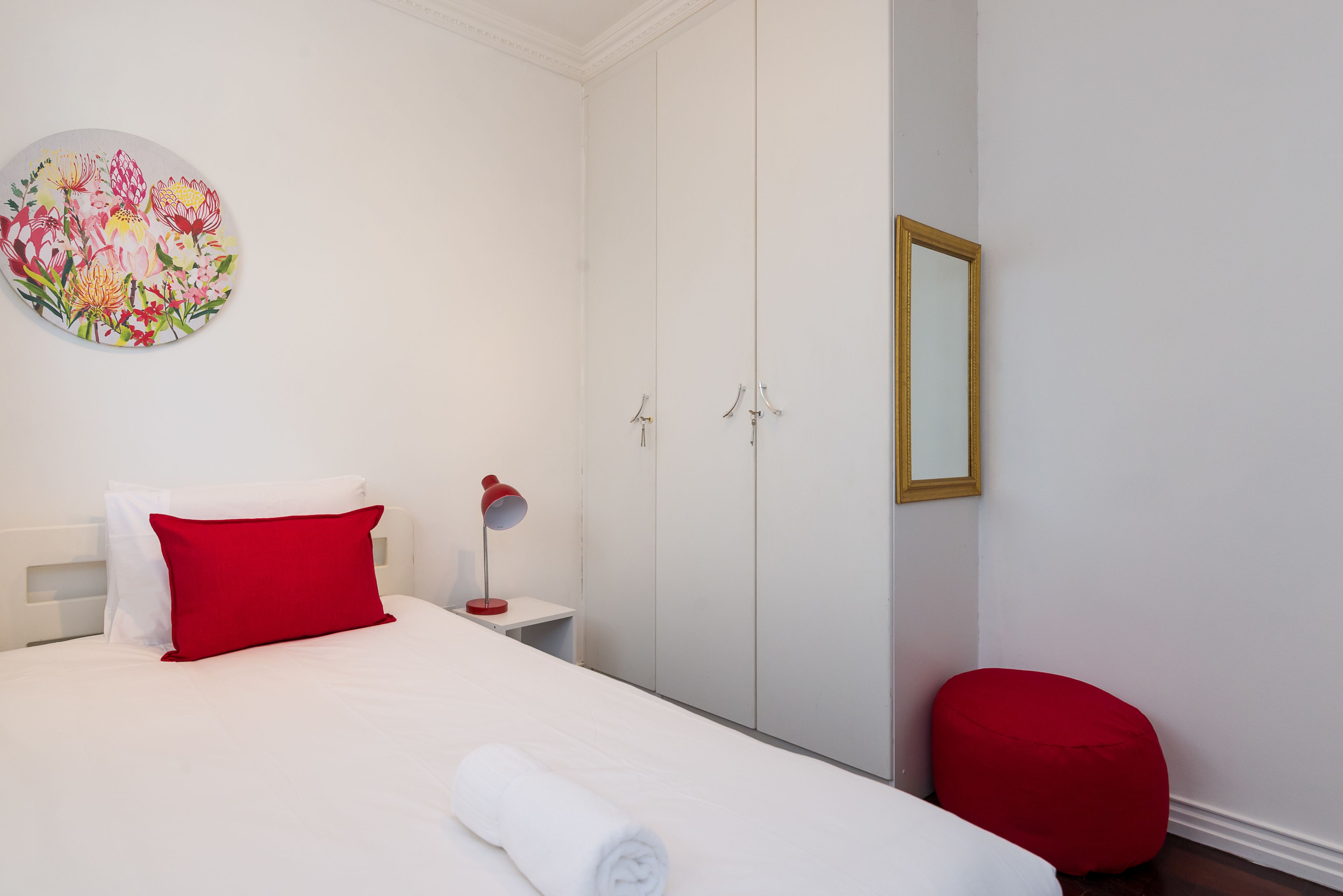 Ein Hotelzimmer mit einem Bett, einem roten Kopfkissen, einer weißen Decke, einer roten Lampe, einem Spiegel und einem roten Hocker.