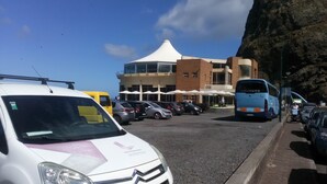 A parking lot with various vehicles, a building with a white roof, and a rocky cliff in the background. - HOUSE CARVOEIRO, SEA AND MOUNTAIN VIEW! (São Vicente, Madeira)