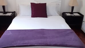A neatly made bed with a white and purple bedspread, a single maroon pillow, and a black bench at the foot of the bed. There are two bedside tables with lamps, and a framed picture hangs above the bed. - HOUSE CARVOEIRO, SEA AND MOUNTAIN VIEW! (São Vicente, Madeira)