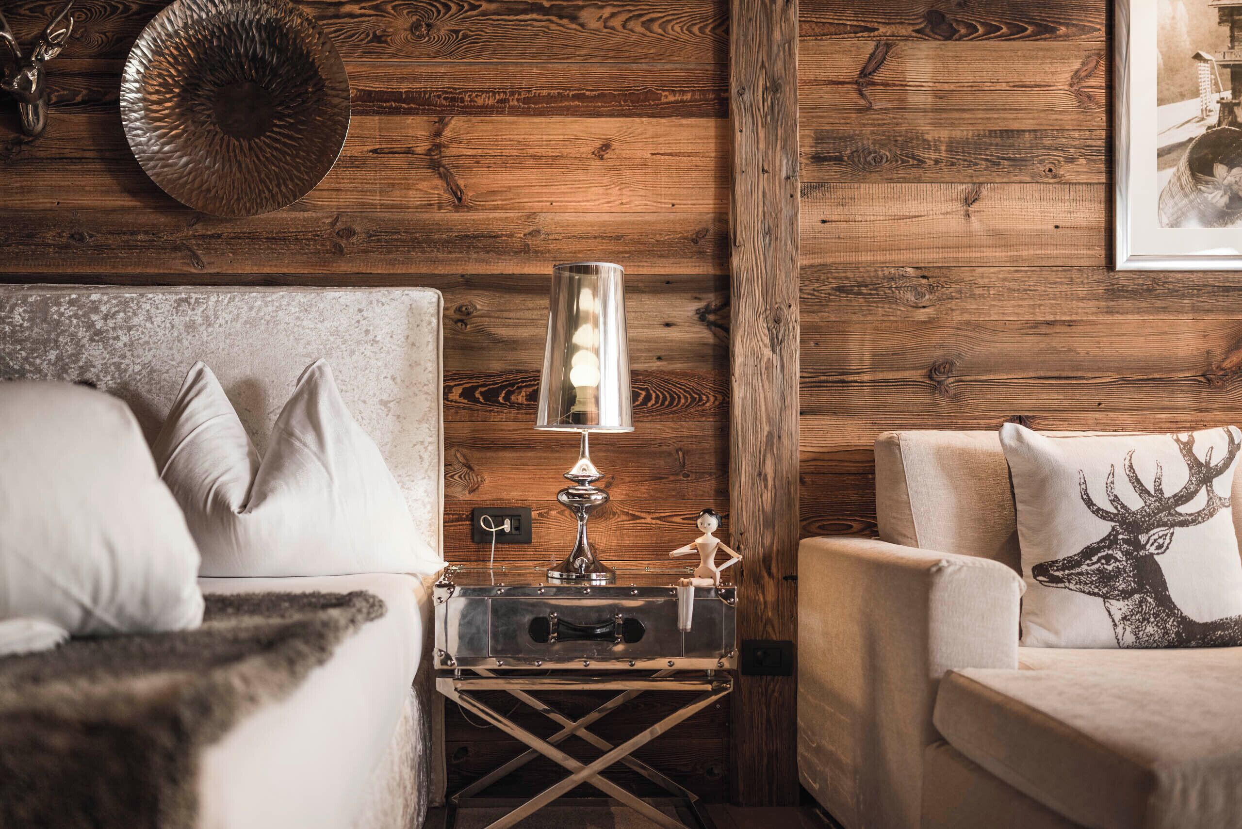 Una habitación acogedora con pared de madera, una cama con ropa blanca, una mesita de noche con lámpara y una silla con un cojín de ciervo.