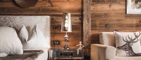 Una habitación acogedora con pared de madera, una cama con ropa blanca, una mesita de noche con lámpara y una silla con un cojín de ciervo.