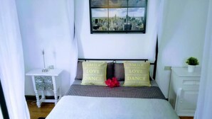 Un dormitorio con cama, mesita de noche, cómoda, ventana y vista panorámica de la ciudad, además de almohadones decorativos.
