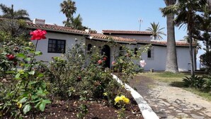 Un edificio blanco con techo de tejas, rodeado por un jardín con diversas plantas y flores.