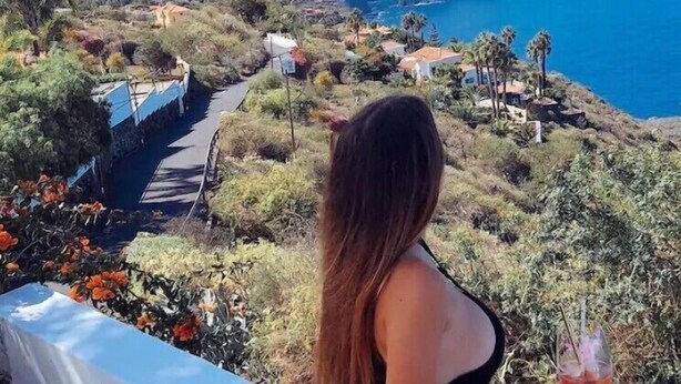 Una persona sentada en una baranda blanca, con vistas a un paisaje costero y una montaña al fondo.