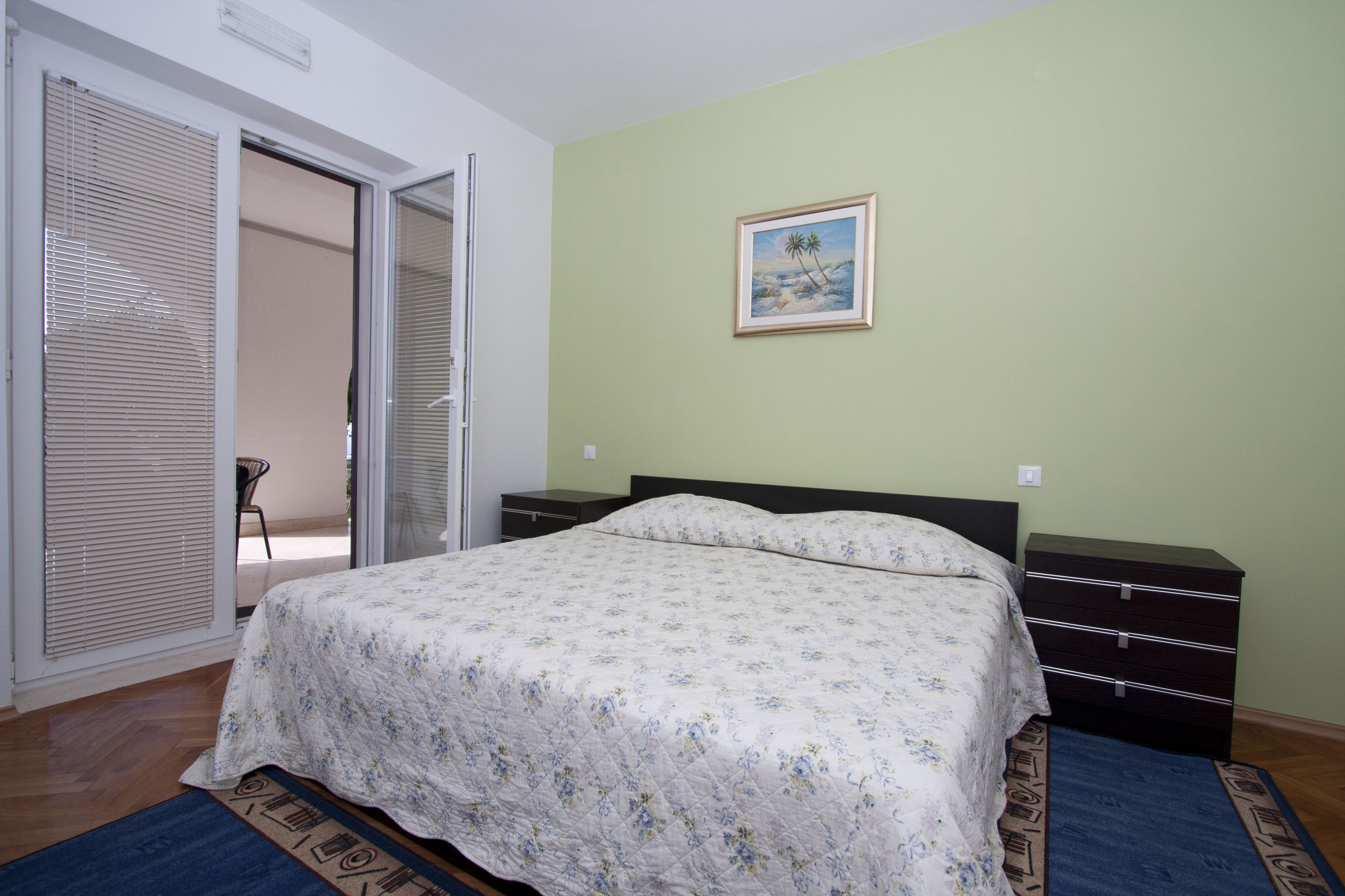 A bedroom with a bed, a nightstand, a dresser, a window with blinds, and a painting on the wall.