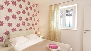 Ein ordentlich bezogenes Bett mit einem rosa Handtuch, ein Nachttisch mit Lampe und ein Fenster mit Meerblick.