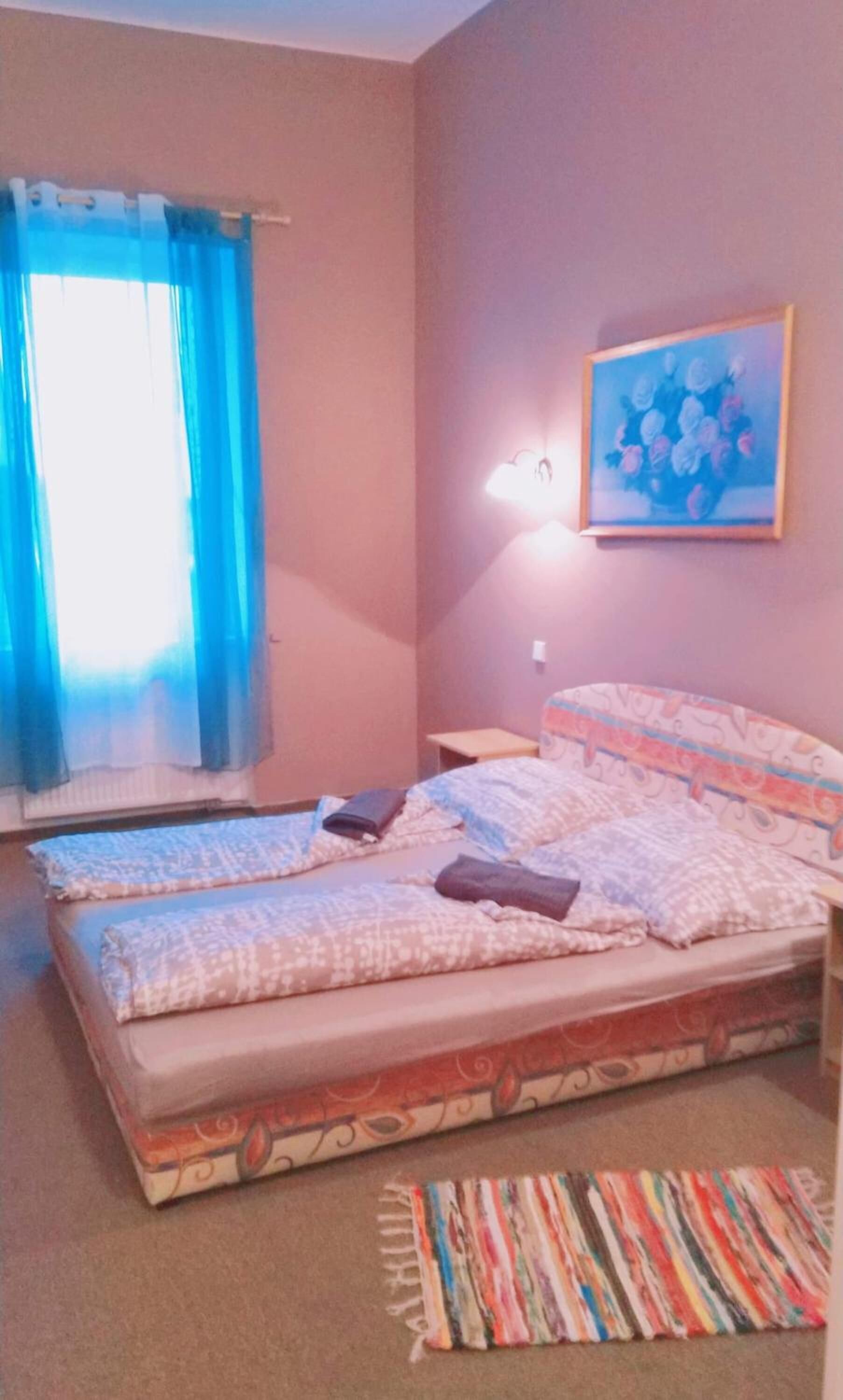 A bedroom with two beds, a window with blue curtains, a painting on the wall, and a colorful rug.