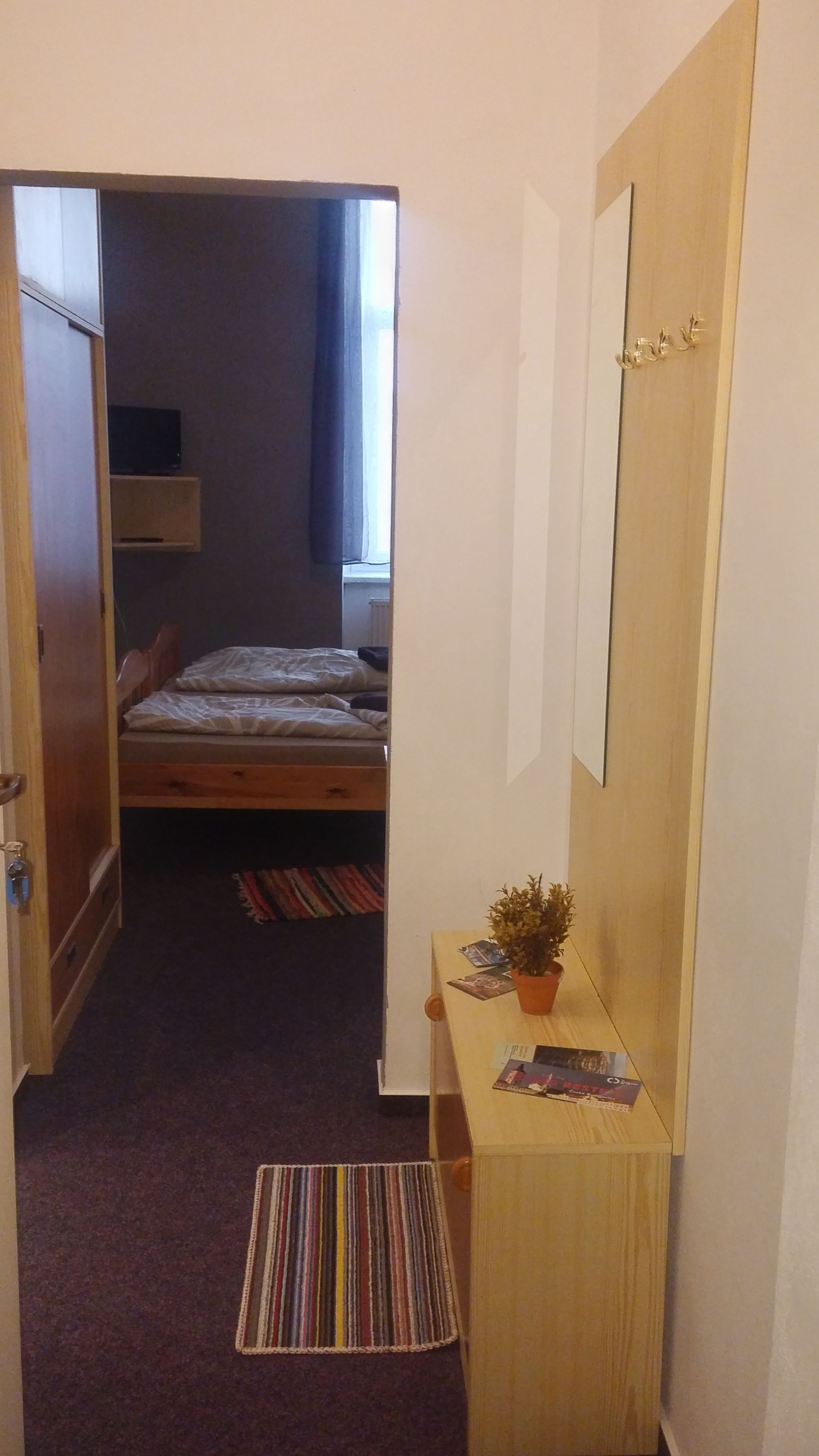 Corredor estreito que leva a um quarto com uma cama, uma pequena escrivaninha com uma planta e uma janela com cortinas.