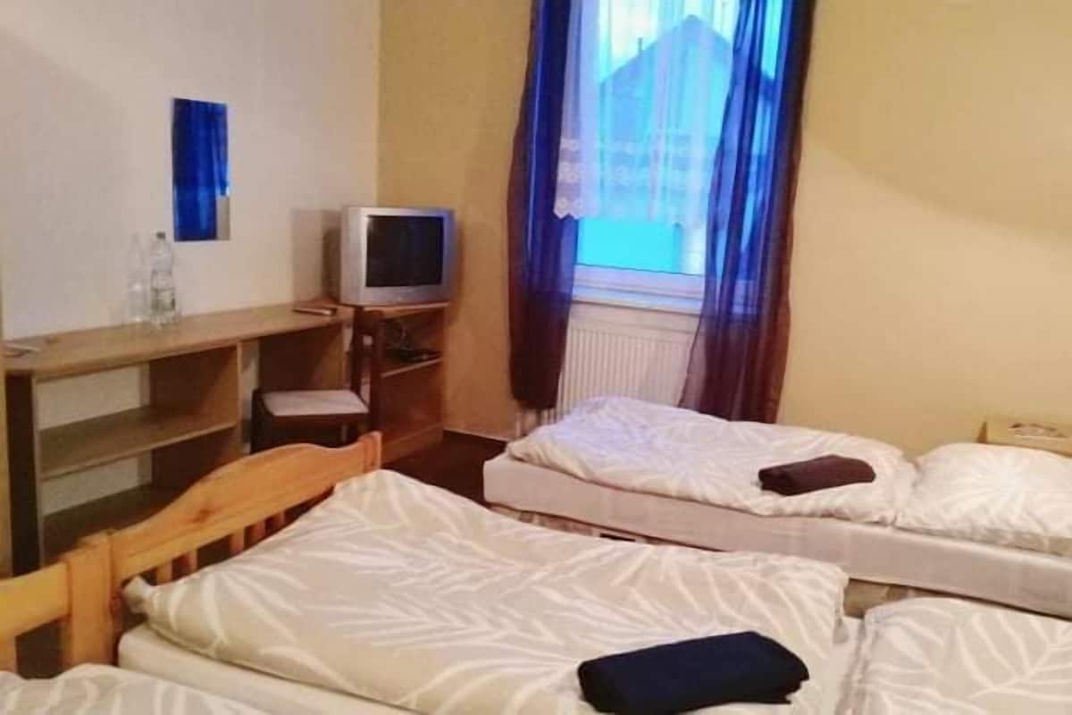 Quarto de hotel pequeno com duas camas de solteiro, uma escrivaninha de madeira com televisão e uma janela com cortinas.