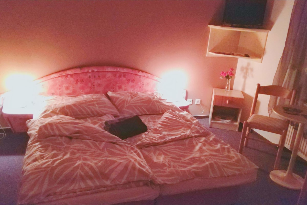 Quarto de hotel com uma cama, mesas de cabeceira, uma cadeira, uma escrivaninha e uma televisão na parede.