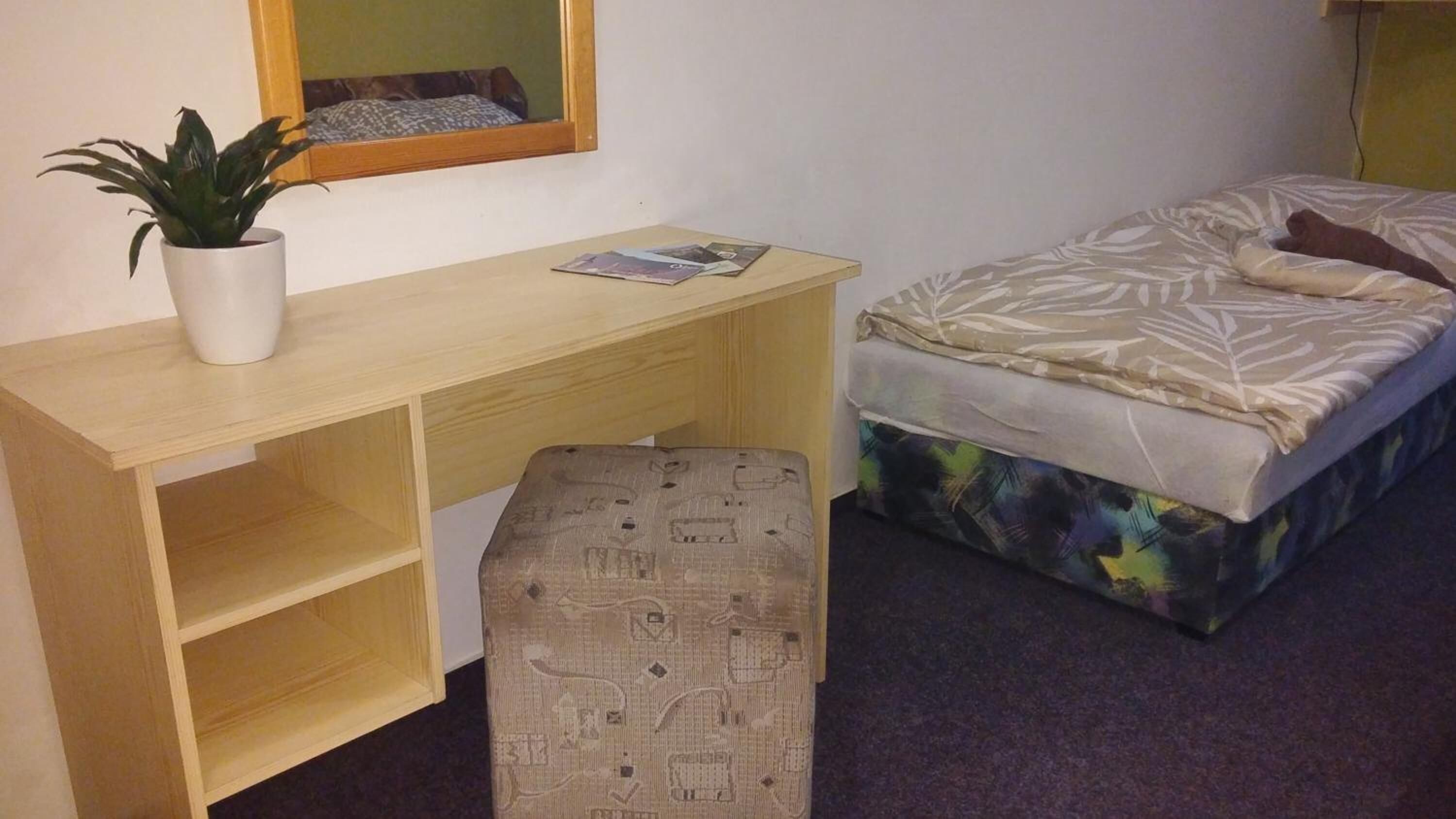 Une petite pièce, de taille modeste, comprenant un bureau en bois, un lit simple, un petit tabouret et une plante en pot.