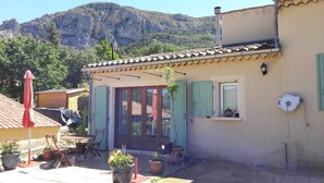 Un pequeño edificio con techo de tejas, puerta con paneles de vidrio y ventana con una pequeña mesa y una tetera. Hay plantas en macetas y un paraguas rojo en el exterior.