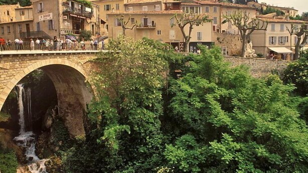 Un puente de piedra con cascada, rodeado de exuberante vegetación y edificios tradicionales.