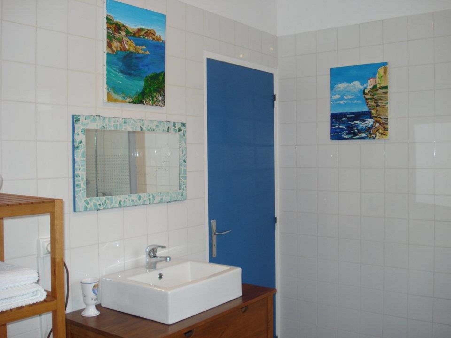 A bathroom with a white sink, a mirror with a mosaic frame, and two paintings of coastal scenes above the door.