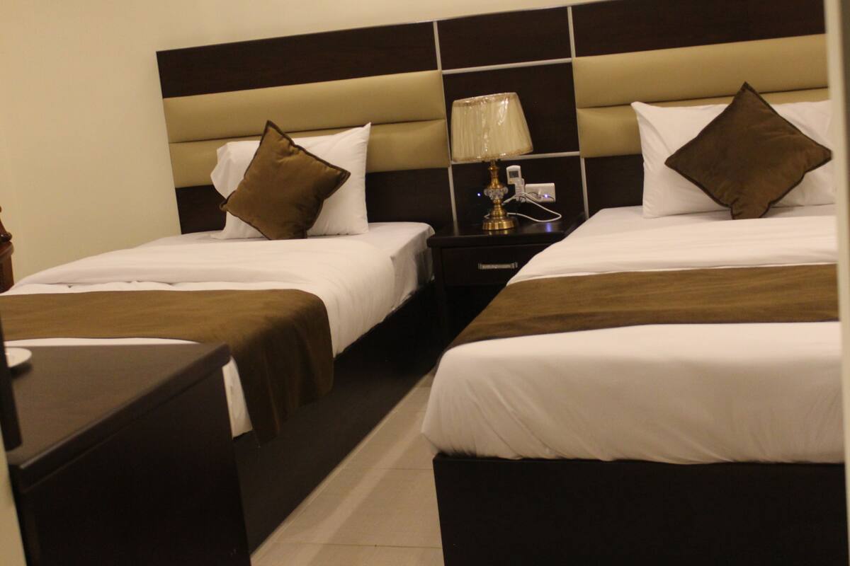 A hotel room with two beds, each with white bedding and brown pillows. There is a nightstand with a lamp between the beds.