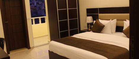 Habitación de hotel con una cama grande, un armario, una mesita de noche con lámpara y vistas a la ciudad a través de una ventana.