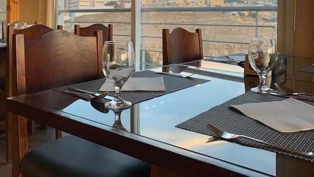 A dining area with a table set for two, large windows offering a city view, and wooden ceiling beams. - Shaqilath Hotel (Wadi Musa)