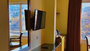 A hotel room with a large window, a flat-screen TV mounted on the wall, a wooden bench, a small table, and a curtain. - Shaqilath Hotel (Wadi Musa)