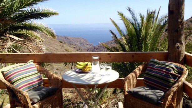 A wooden deck with wicker chairs and a small table, overlooking a scenic view of the ocean and hills. - CASA ALMA a dream look that makes you happy (Hermigua) (Hermigua)