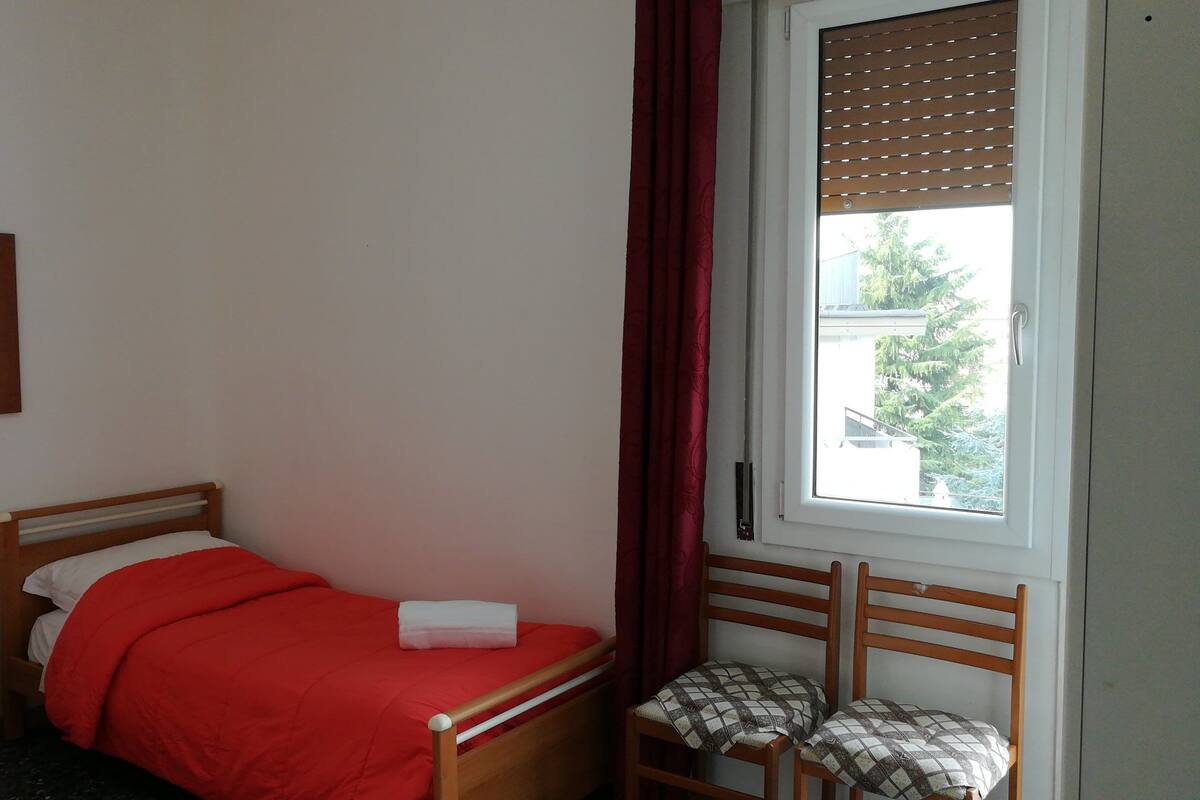 A small bedroom with a single bed, a bedside table with a remote control, a window with blinds, and a mirror on the wall.
