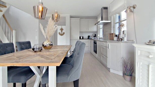 A modern kitchen with white cabinets, a wooden dining table, and grey upholstered chairs. - Living under thatch in Seasons - fireplace & sauna  (Groß Schwansee)
